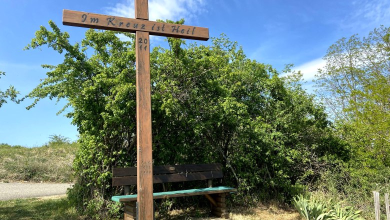 Holzkreuz mit Inschrift 'Im Kreuz ist Heil' und Sitzbank, umgeben von Büschen und blauem Himmel.