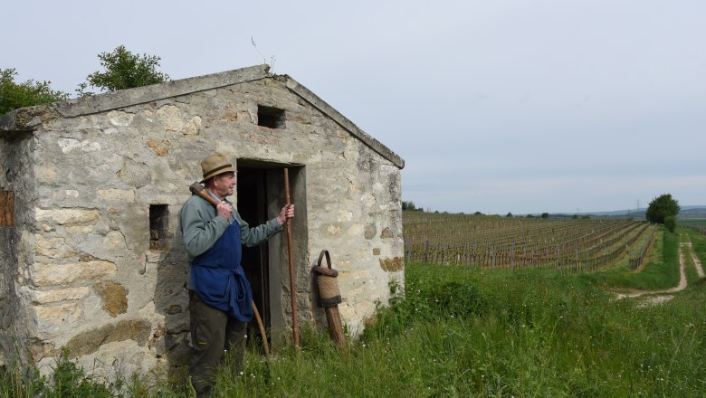 Ein Mann mit Strohhut steht vor einer kleinen Steinhütte in einem Weinberg.