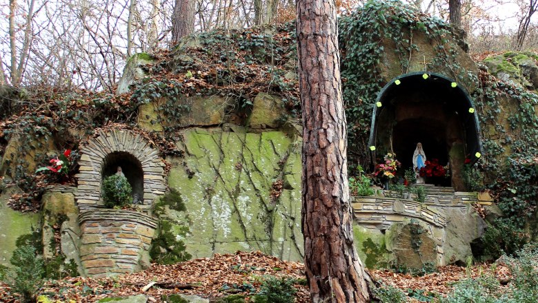 Eine steinerne Grotte mit einer Marienstatue und Pflanzen, umgeben von Bäumen und Laub.