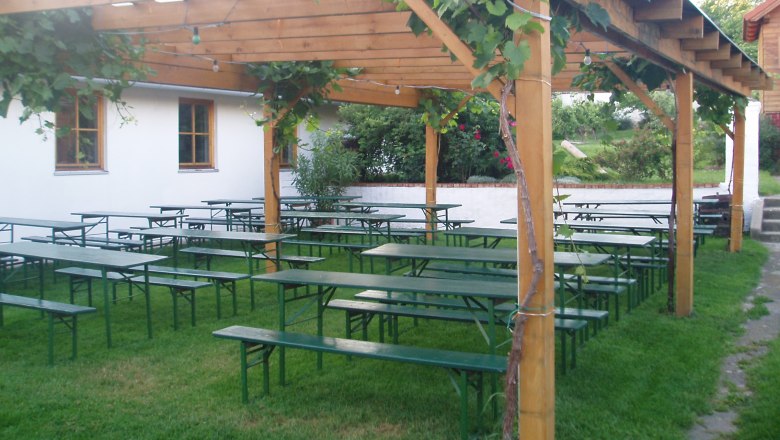 Ein Biergarten mit Holzb&auml;nken und Tischen unter einer Pergola mit Weinreben.