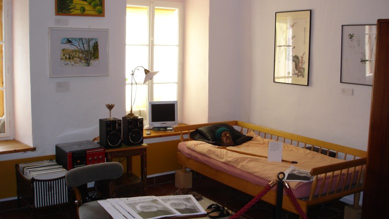 Innenansicht eines Museumszimmers mit Bett, Bildern an der Wand und einem kleinen Schreibtisch mit Computer.