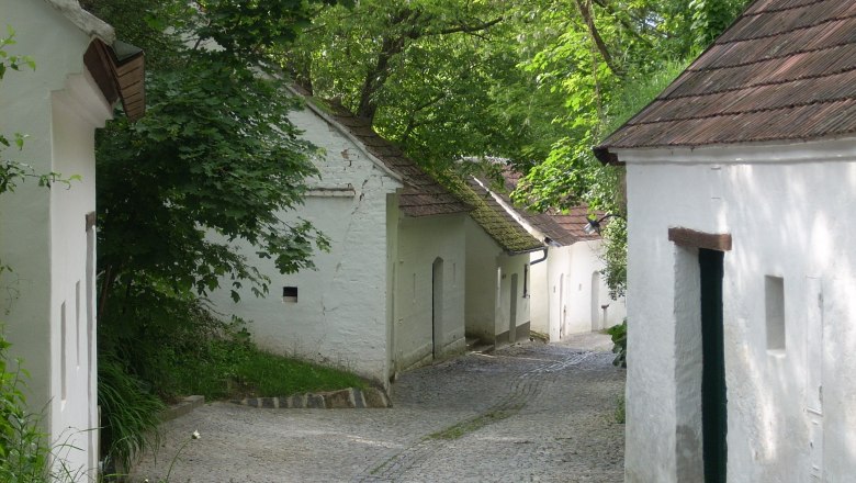 Eine malerische Kellergasse mit wei&szlig;en Weinkellern und Kopfsteinpflaster, umgeben von gr&uuml;nen B&auml;umen.