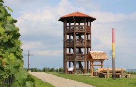 Ein hölzerner Aussichtsturm mit rotem Dach steht auf einem Hügel neben einem Kreuz und einer Sitzbank. Der Himmel ist bewölkt.