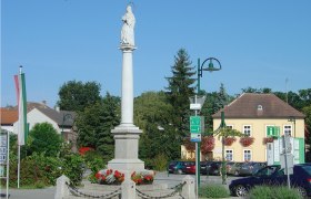 Statue auf einer Säule in einem Platz in Haugsdorf, umgeben von Gebäuden und Bäumen.