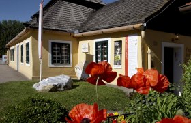 Gelbes Haus mit Schindeldach und Mohnblumen im Vordergrund.
