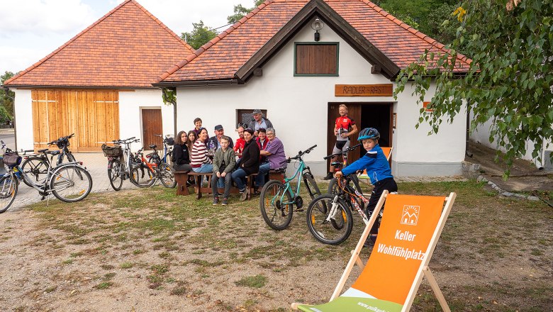 Gruppe von Menschen sitzt vor einem Gebäude mit Fahrrädern, ein Kind steht mit Fahrrad daneben. Ein Liegestuhl mit Aufschrift ist im Vordergrund.