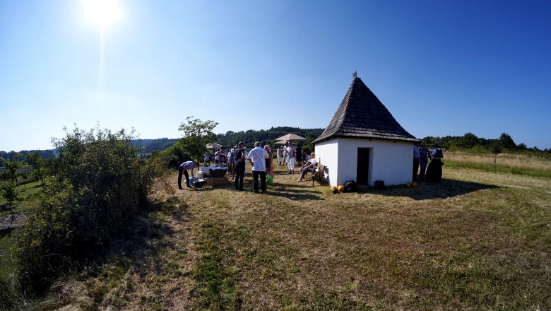 Eine Gruppe von Menschen versammelt sich bei einer kleinen, weißen Hütte mit spitzem Dach auf einer Wiese unter klarem Himmel.
