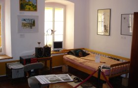 Innenansicht eines Museumszimmers mit Bett, Bildern an der Wand und einem kleinen Schreibtisch mit Computer.