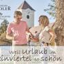 Ein Mann und eine Frau stehen mit Weingläsern und Wanderstöcken vor einem Baum, im Hintergrund ein Gebäude mit Turm.
