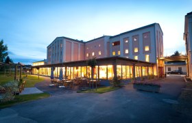 Abendaufnahme eines beleuchteten Geb&auml;udes mit Terrasse und Gartenm&ouml;beln.