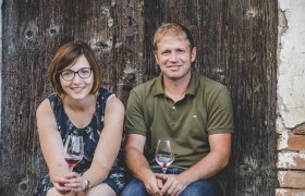 Ein Mann und eine Frau sitzen vor einer Holzwand und halten Weingläser in den Händen.