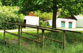 Eine kleine Holzbrücke führt über eine Wiese zu weißen Gebäuden mit grünen Türen, umgeben von Bäumen und Büschen.