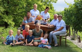Eine Familie sitzt und steht um einen Tisch im Freien, umgeben von B&auml;umen und Gras.