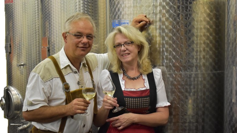 Ein Mann und eine Frau in traditioneller Kleidung stehen vor Weintanks und halten Weingläser.