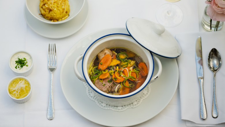 Ein gedeckter Tisch mit einem Eintopf in einem weißen Topf, einem Knödel, zwei Soßen und einem Glas Weißwein.