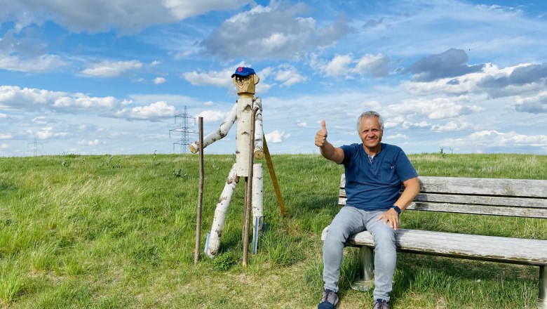 Ein Mann sitzt auf einer Bank neben einer Holzfigur auf einem Feld unter blauem Himmel.