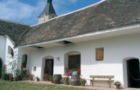 Traditionelles Gebäude mit weißer Fassade, Holztüren und Blumen in Töpfen. Im Hintergrund ein Turm mit Uhr.