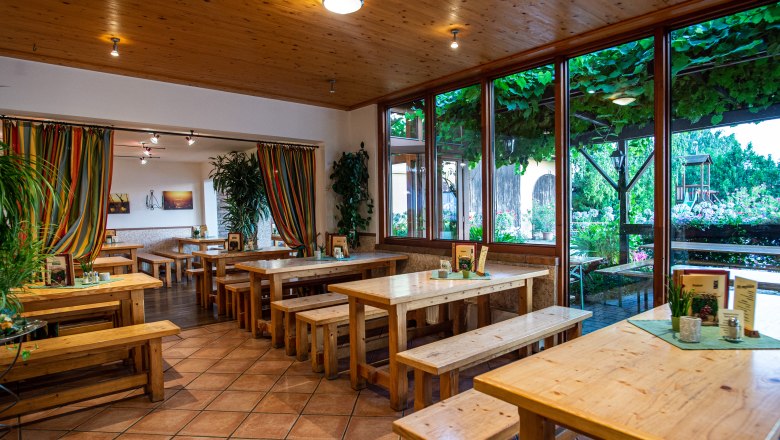 Innenansicht eines Heurigenlokals mit Holztischen und Bänken, bunten Vorhängen und großen Fenstern mit Blick auf einen begrünten Außenbereich.