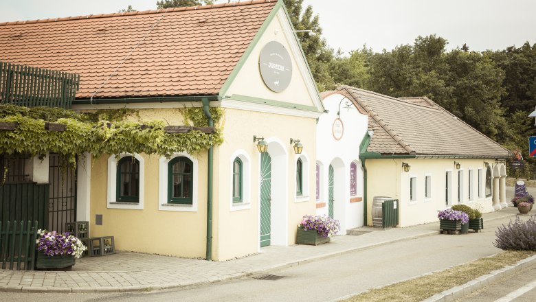 Außenansicht eines traditionellen Heurigen mit gelber Fassade und roten Dachziegeln, umgeben von Pflanzen.