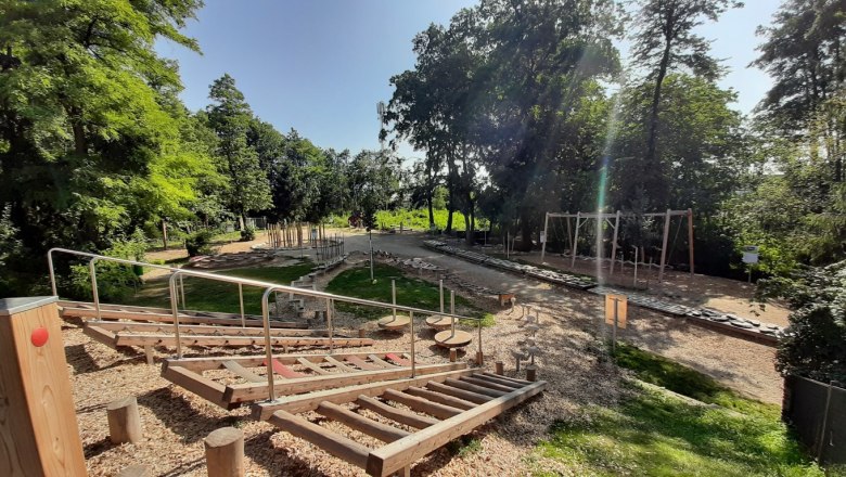 Ein Motorikpark mit verschiedenen Holzelementen und Kletterger&auml;ten, umgeben von B&auml;umen und Gr&uuml;nfl&auml;chen.