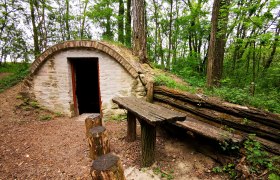Steinh&uuml;tte im Wald mit Holztisch und Baumst&uuml;mpfen als Sitzgelegenheiten.