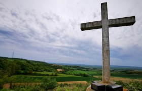 Ein gro&szlig;es Steinkreuz auf einem H&uuml;gel mit Blick auf eine gr&uuml;ne Landschaft und bew&ouml;lkten Himmel.