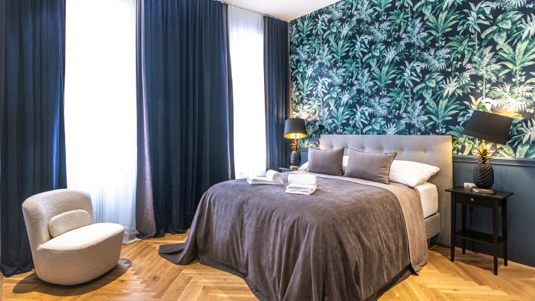 Modernes Doppelzimmer mit grauem Bett, tropischer Tapete und dunklen Vorhängen.