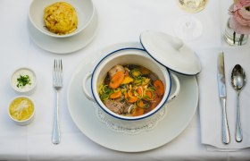Ein gedeckter Tisch mit einem Eintopf in einem weißen Topf, einem Knödel, zwei Soßen und einem Glas Weißwein.