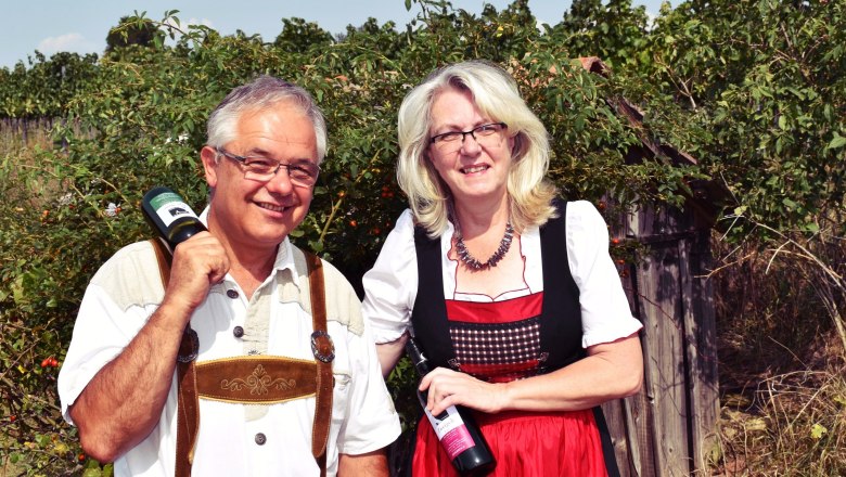 Ein Mann in Lederhosen und eine Frau im Dirndl halten Weinflaschen in einem Weinberg.