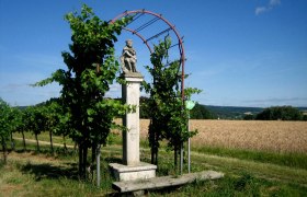 Steinernes Kreuz mit Figur in ländlicher Umgebung, umgeben von Pflanzen und Feldern.