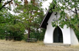 Kleine weiße Kapelle im Wald mit Holzbänken davor.