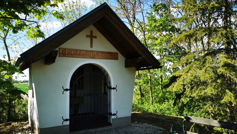 Kleine Kapelle im Grünen mit Holzkreuz und Bank daneben.