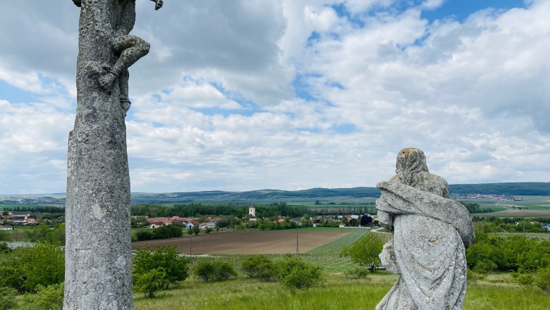 Zwei Steinskulpturen auf einem H&uuml;gel mit Blick auf eine l&auml;ndliche Landschaft und ein Dorf im Hintergrund.
