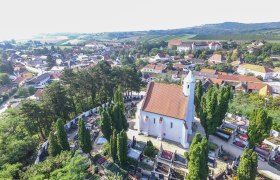 Luftaufnahme einer Kirche mit rotem Dach, umgeben von einem Friedhof und Bäumen, in einer ländlichen Umgebung mit Häusern und Hügeln im Hintergrund.