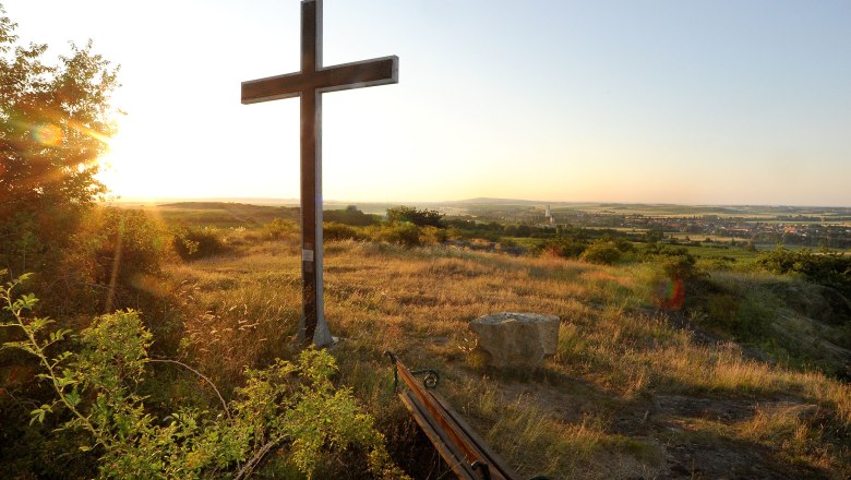 Ein Kreuz auf einem H&uuml;gel bei Sonnenuntergang mit Blick auf eine weite Landschaft und ein Dorf im Hintergrund.