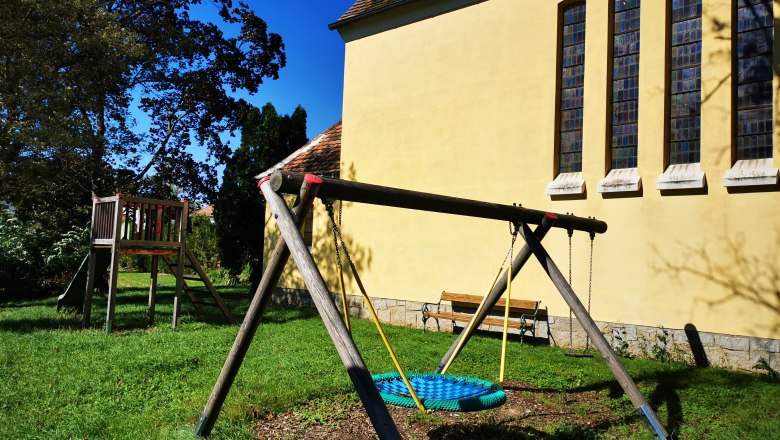 Spielplatz mit Schaukel und Rutsche neben einer gelben Kapelle mit hohen Fenstern.