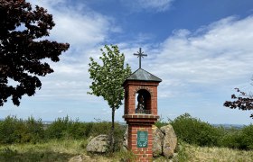 Ein gemauerter Bildstock mit Kreuz auf einem Hügel, umgeben von Bäumen und Sträuchern, unter einem blauen Himmel.