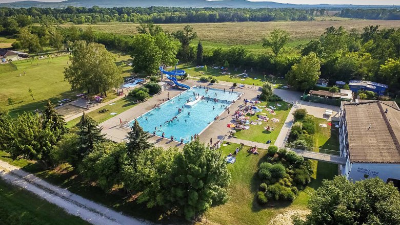 Luftaufnahme eines Freibads mit Schwimmbecken, Rutsche und Liegewiese, umgeben von Bäumen und Feldern.