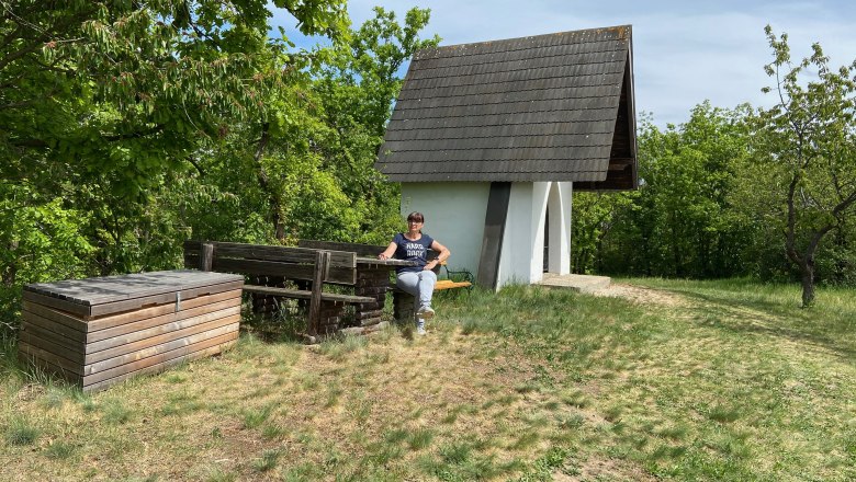 Eine kleine wei&szlig;e Kapelle mit spitzem Dach in einer gr&uuml;nen Landschaft, daneben eine Person auf einer Bank.