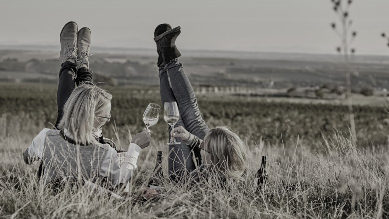 Zwei Personen liegen im Gras mit Weingläsern in der Hand, die Beine in die Luft gestreckt, vor einer weiten Landschaft.