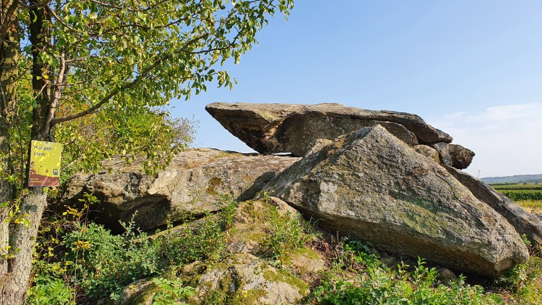 Große Felsformation mit Baum und Schild im Vordergrund, blauer Himmel im Hintergrund.