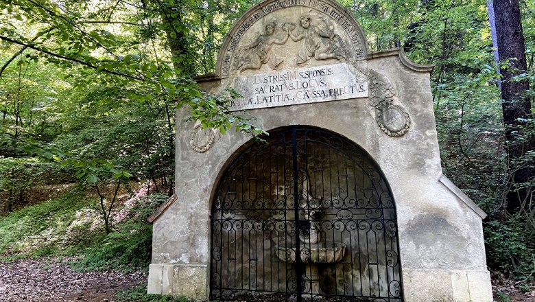 Historische Brunnstube im Wald mit verziertem Tor und lateinischer Inschrift.