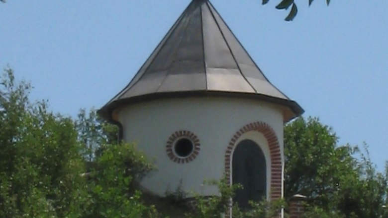 Ein runder Wasserturm mit spitzem Dach und Wetterfahne, umgeben von Bäumen und Wiese.