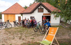 Gruppe von Menschen sitzt vor einem Gebäude mit Fahrrädern, ein Kind steht mit Fahrrad daneben. Ein Liegestuhl mit Aufschrift ist im Vordergrund.
