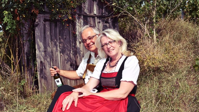 Ein Paar in traditioneller Kleidung sitzt vor einer Holzhütte im Freien.