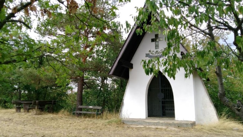 Kleine wei&szlig;e Kapelle im Wald mit Holzb&auml;nken davor.