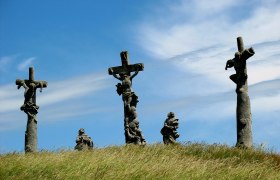 Drei Kreuze mit Figuren auf einem Hügel unter blauem Himmel.