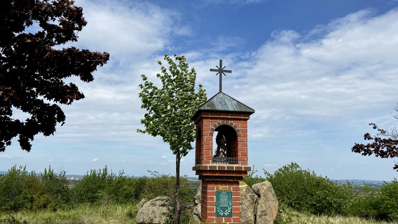 Ein gemauerter Bildstock mit Kreuz auf einem Hügel, umgeben von Bäumen und Sträuchern, unter einem blauen Himmel.