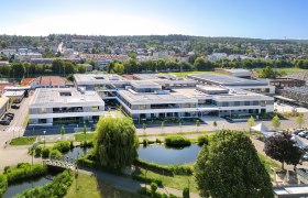 Luftaufnahme eines modernen Schulcampus mit Solarpanelen auf den D&auml;chern, umgeben von Gr&uuml;nfl&auml;chen und einem Teich im Vordergrund.