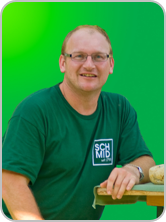 Person in grünem T-Shirt mit Logo, lächelnd vor grünem Hintergrund.